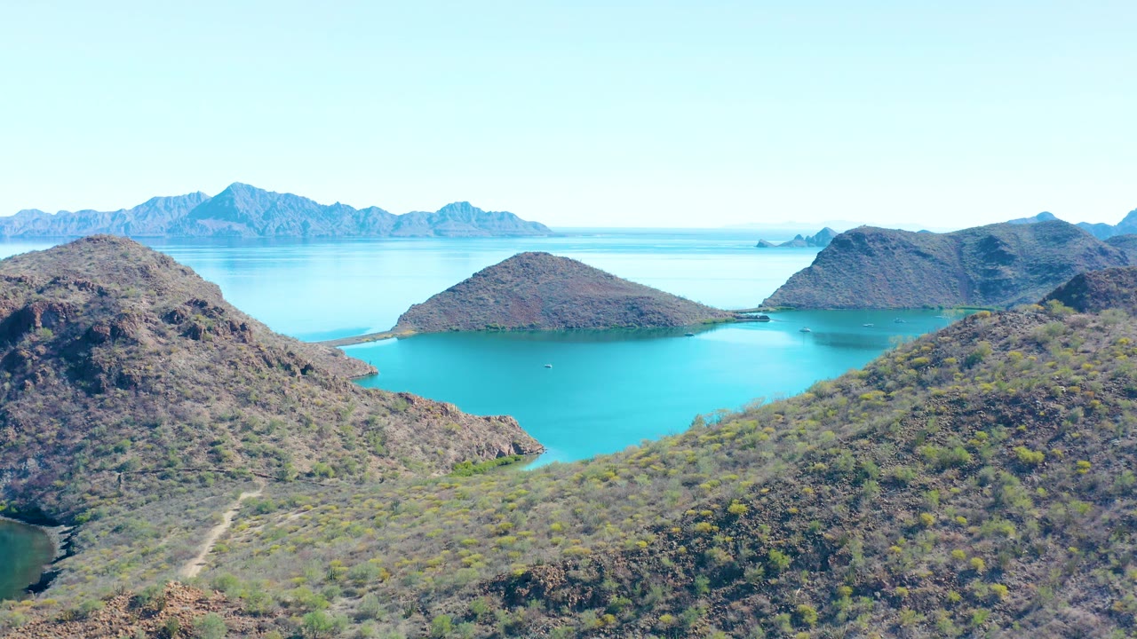 A high aerial frame over Juncalito shows dry, brushy hills framing bright blue water, with a small boat faintly visible on the bay and no visible people.