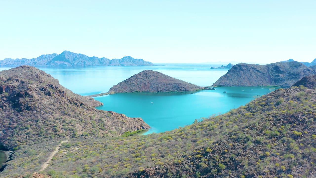 A wide drone-style frame shows Juncalito's blue bay enclosed by rocky hills, a tiny boat on the water, pale sky, and no visible people.