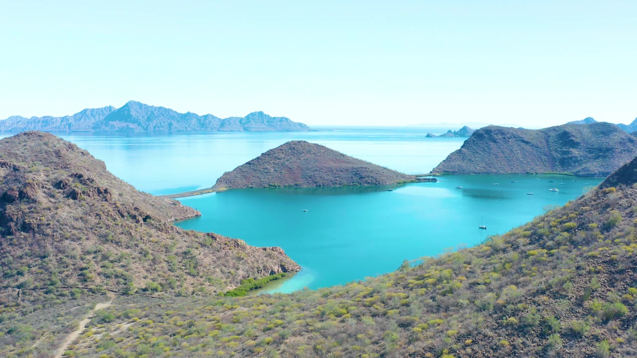 Aerial frame over Juncalito showing a rounded rocky hill, bright blue-green bay water, small boats in the distance, and no visible people.