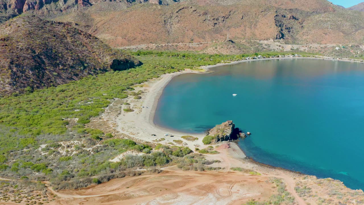 Aerial frame over Juncalito with the pale beach curving around clear blue-green water, a white boat near the center of the cove, rocky hills behind, and no visible people.