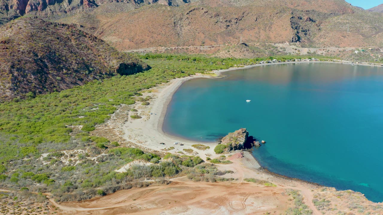 Aerial view over Juncalito showing the sandy curve of the cove, a small boat on calm turquoise water, dense green vegetation along shore, rocky hills, and no visible people.