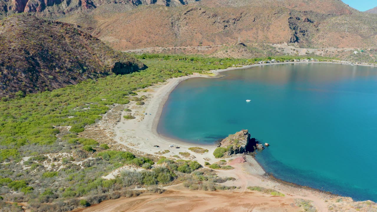 Aerial frame of Juncalito, Baja California Sur showing a curved sandy beach, turquoise bay water, green scrub, arid hills, a rocky point, and a small white boat offshore.