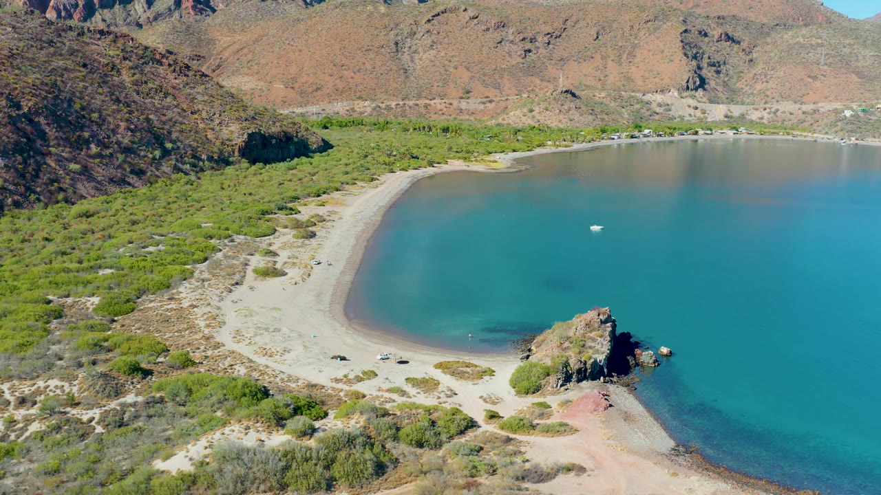 High drone frame over Juncalito with the beach arc centered, low green vegetation along the shore, several small vehicles near the sand track, a white boat in the bay, and no visible people.