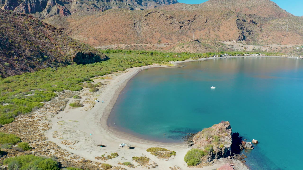 Aerial frame over Juncalito, Baja California Sur, showing a curved sandy beach, turquoise bay water, scrub vegetation, parked cars, rocky hills, and no visible people at the start of the clip.