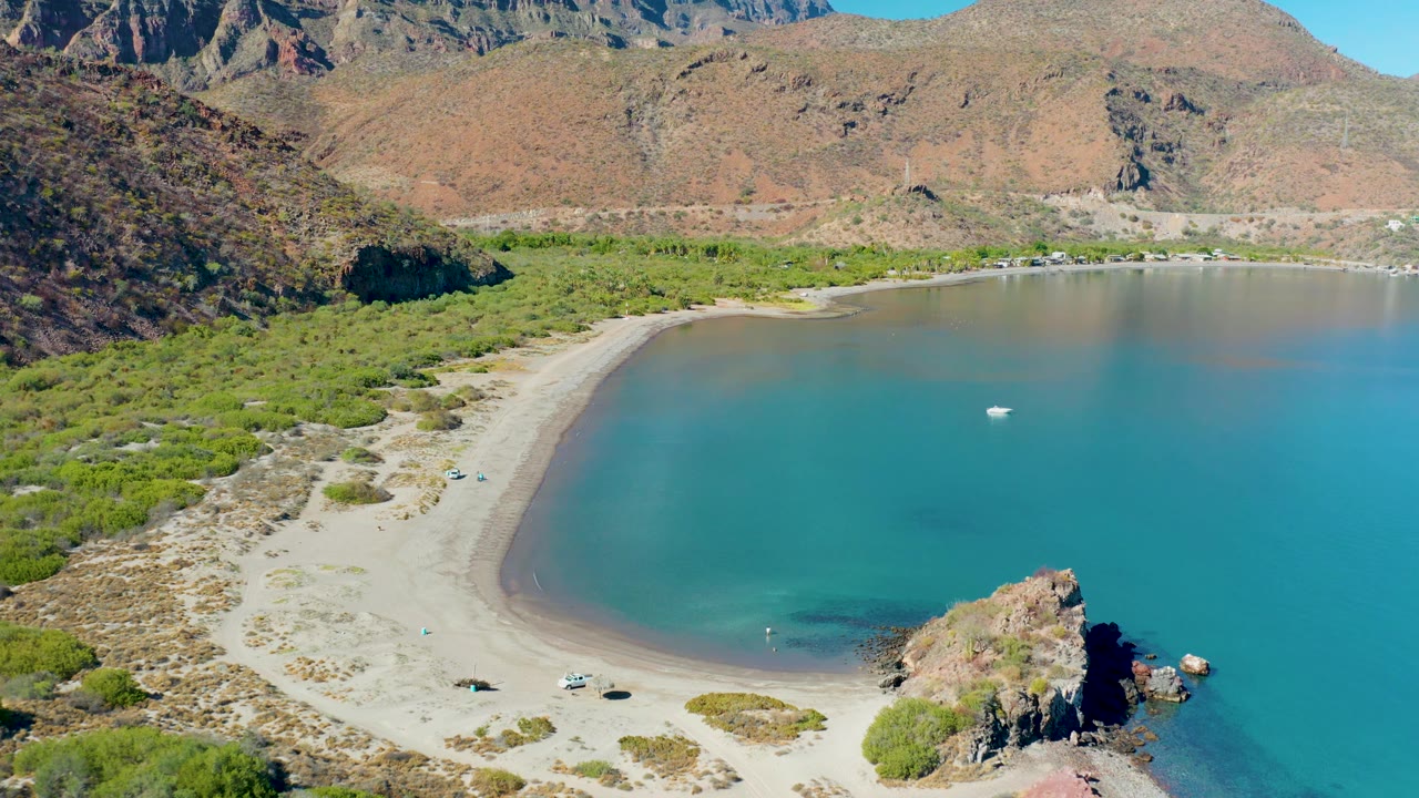 Curved shoreline with shallow turquoise water, beach cars, and hills behind.