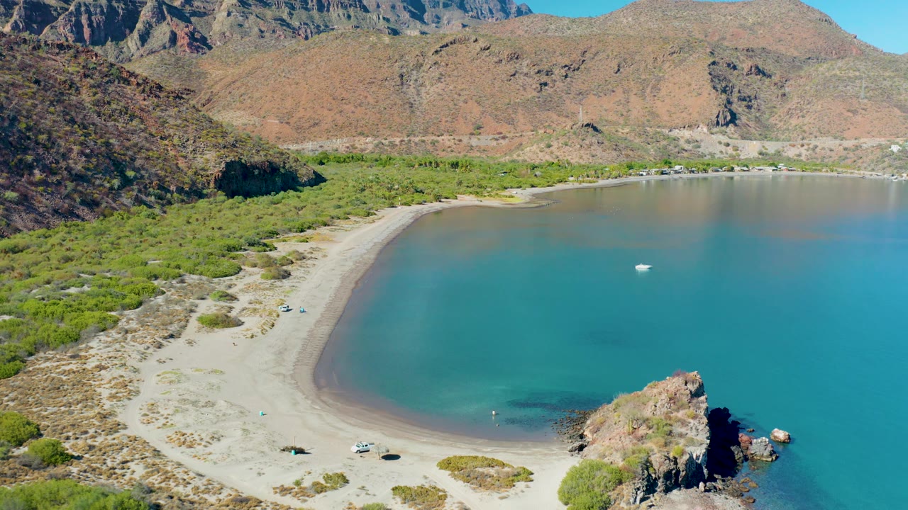 Wide aerial frame over Juncalito showing more open turquoise water to the right, the curved beach below the hills, a rocky point at lower right, and no visible people.