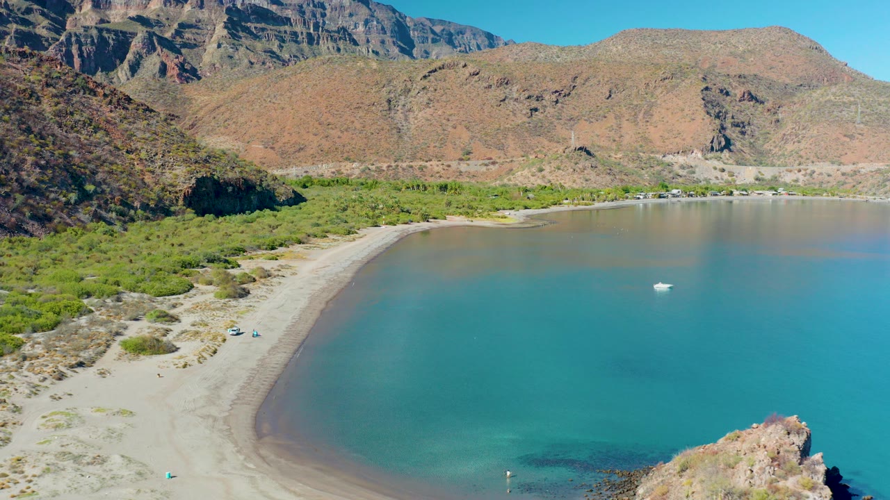 Aerial frame over Juncalito, Baja California Sur showing a curved sandy beach, turquoise bay water, shoreline greenery, rocky mountains, a small boat offshore, and no visible people.