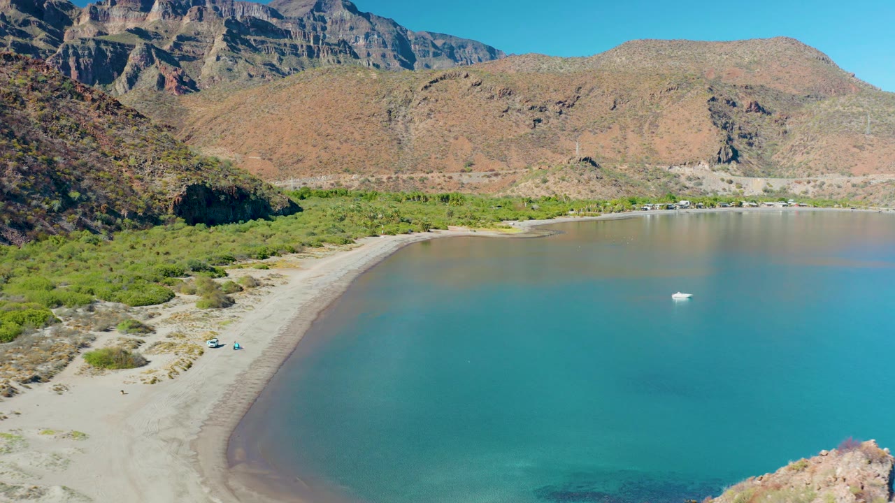 Aerial shot over Juncalito, Baja California Sur showing the beach curve along the left edge, smooth turquoise water across the bay, distant rocky mountains, and no visible people.