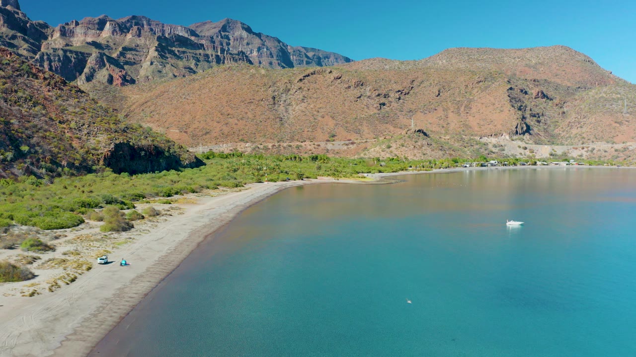 Aerial frame of Juncalito, Baja California Sur, with a curved sandy shore at left, turquoise water at right, a small white boat offshore, rugged mountains behind, and tiny people visible near the lower beach.