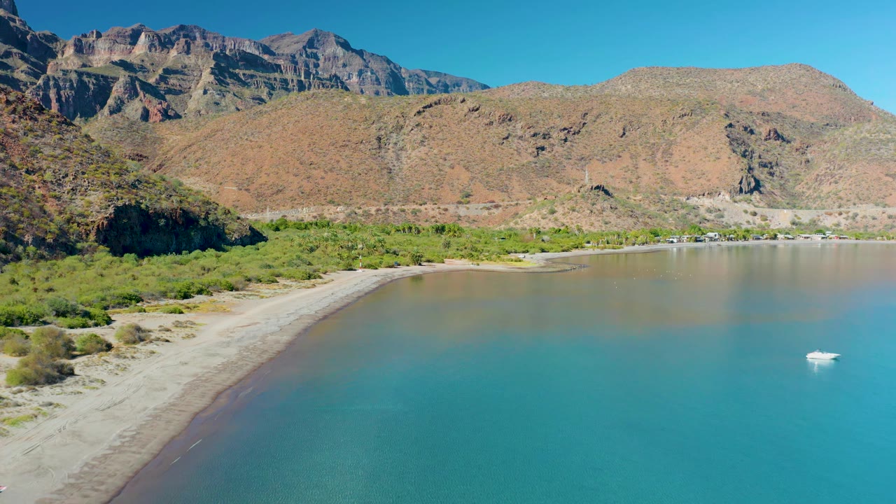 Juncalito aerial shoreline view with pale sand along the left curve, blue-green water across the lower frame, desert mountains behind, and the small white boat closer to the right edge, with no visible people.