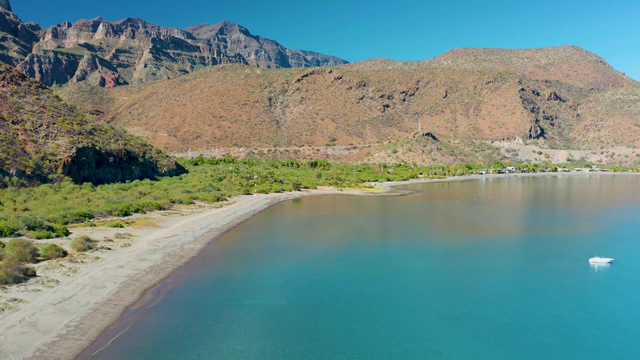 Wide aerial view of Juncalito bay with the sandy shoreline sweeping in from the left, turquoise water filling the foreground, rocky mountains across the back, and the boat near the far right, with no visible people.