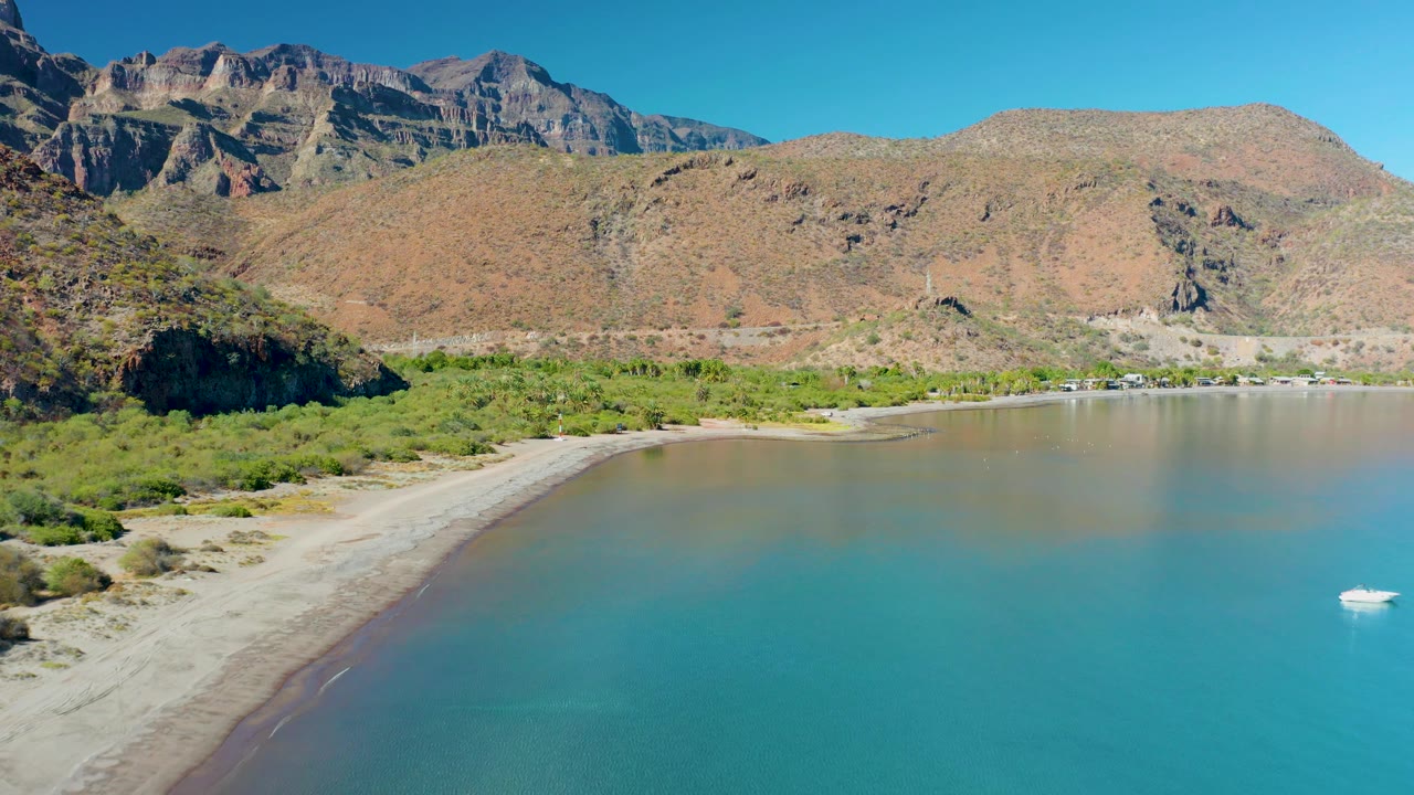 Aerial frame over Juncalito with the beach running diagonally from the lower left, green vegetation at the shore, rocky mountains in bright light, and the white boat low at the right edge, with no visible people.