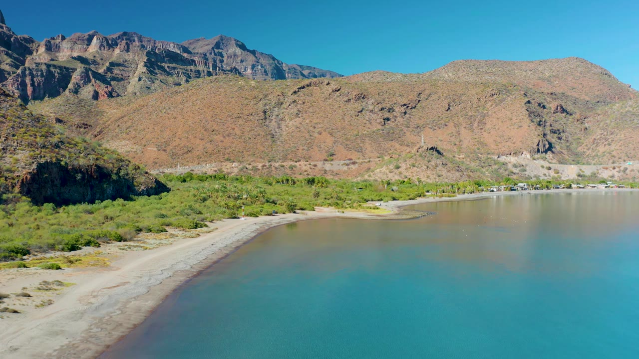 At 0:01, the Juncalito shoreline remains centered with shallow blue-green water, exposed sand along the left curve, brush beyond the beach, mountains, and no visible people.