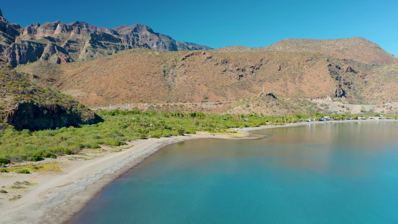 At 0:02, blue-green water fills more of the foreground while the sandy edge arcs along the left side, with Juncalito brush, dry mountain ridges, and no visible people.