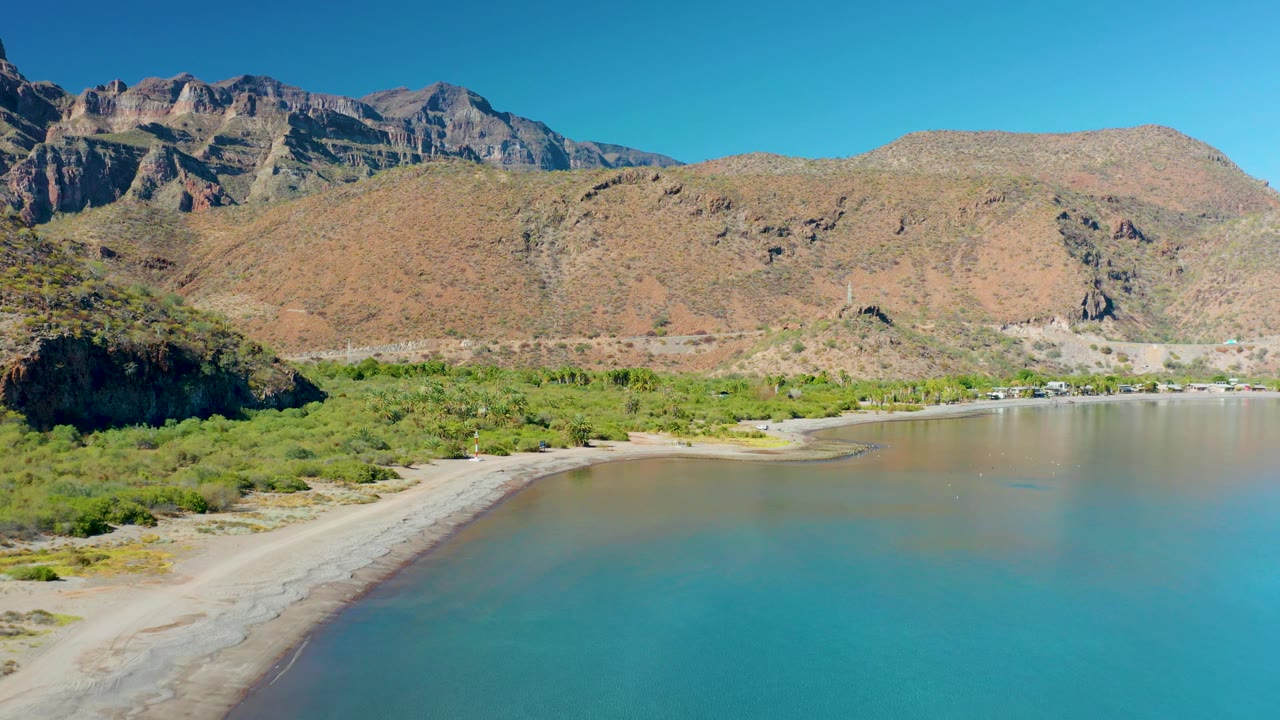 At 0:03, the waterline sits below the sandy strip, with a dark rocky hill at left, green vegetation across the shore, Juncalito mountains behind, and no visible people.