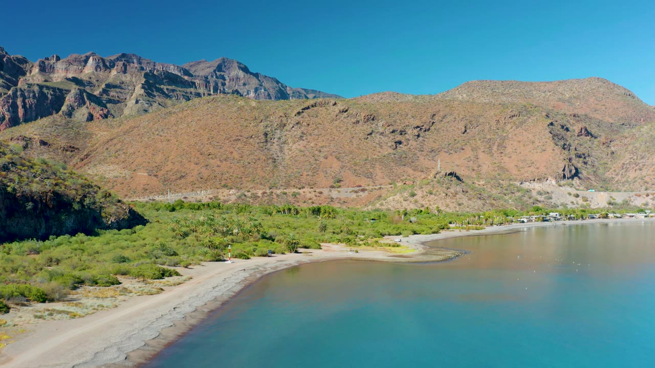 Aerial view over Juncalito with clear turquoise and shallow brown water in front of a sandy beach, green vegetation, rocky mountains, and no visible people.