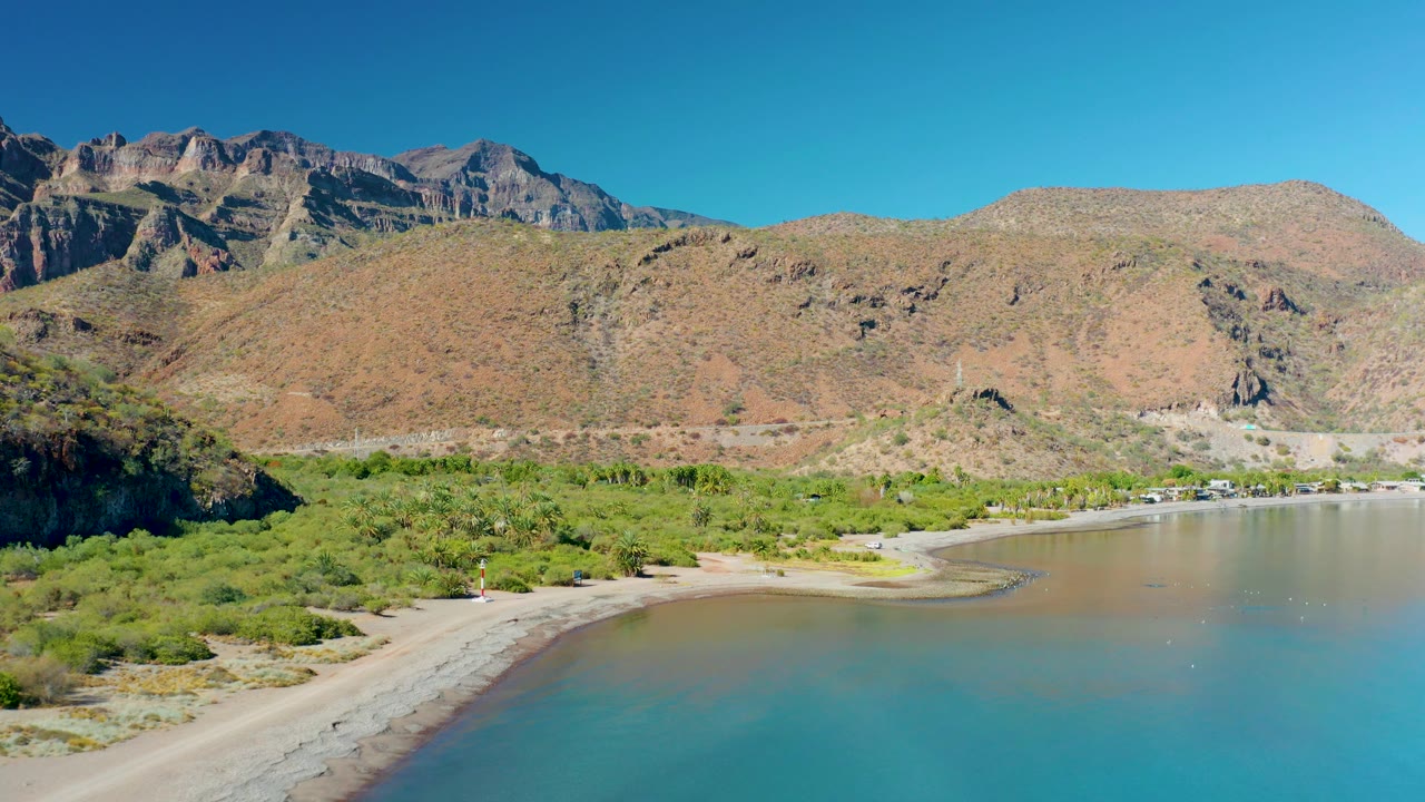 Aerial view of Juncalito shoreline showing a small vehicle on the sandy beach near green vegetation, shallow bay water, rocky mountains, and no visible people.