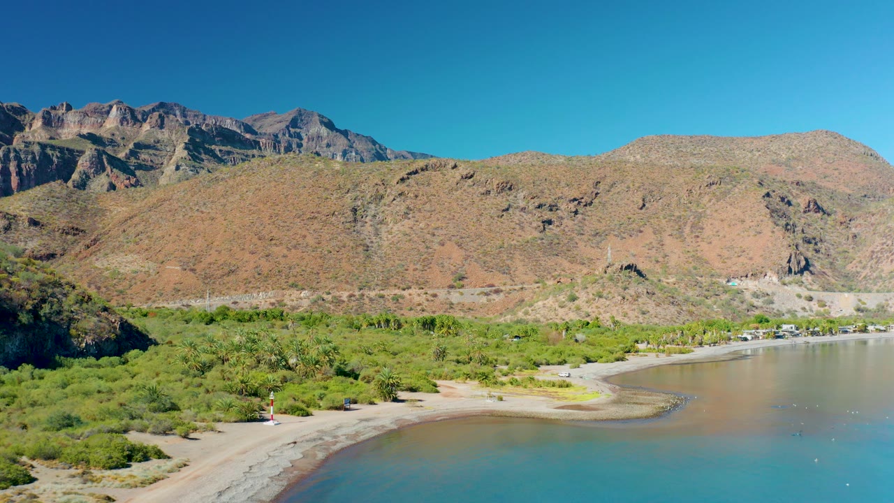 Aerial frame over Juncalito, Baja California Sur, showing a curved sandy shore, green coastal brush, calm blue-green water, tan mountains, clear sky, and no visible people.