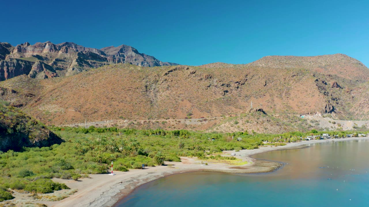 Wide aerial coastline with beach curve, vegetation, road, water, and mountains.