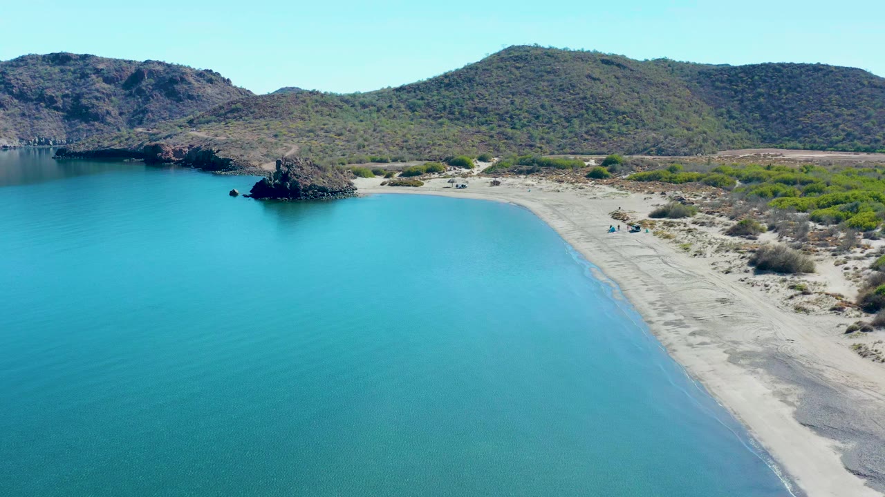 Aerial view in Juncalito with the shoreline curving farther through the frame, clear blue water at left, pale sand at right, dark rocks near the cove, and no visible people.