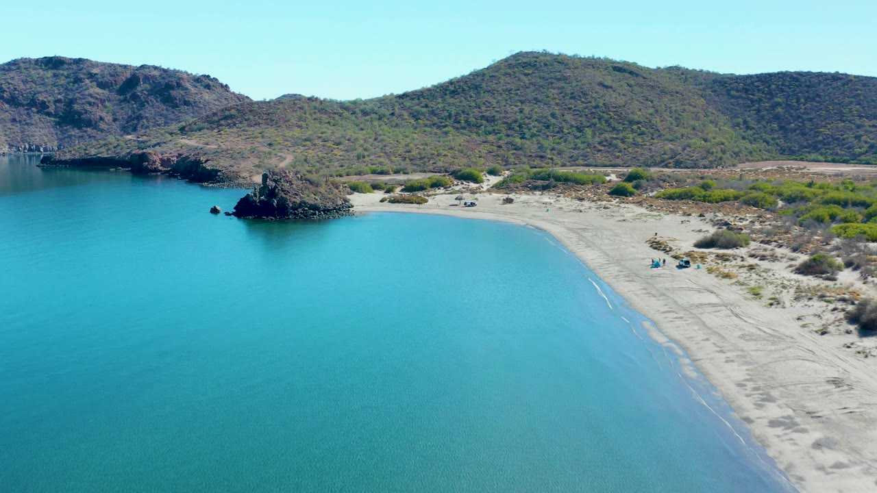 Aerial view of Juncalito, Baja California Sur, with turquoise water filling the foreground, a sandy beach curving to the right, a dark rocky outcrop near shore, dry hills behind, and no visible people.