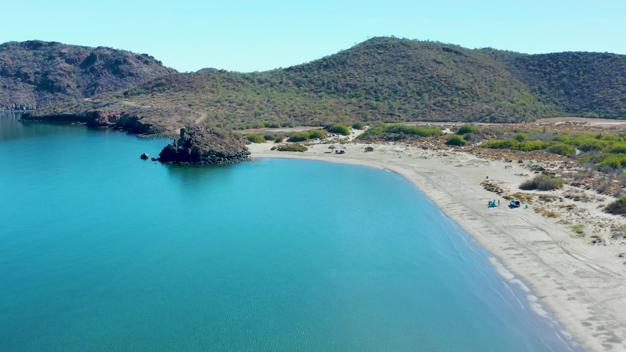 Aerial frame over Juncalito with the rocky outcrop more central, calm turquoise water in front, a pale beach sweeping along the right side, low green shrubs behind the sand, and no visible people.