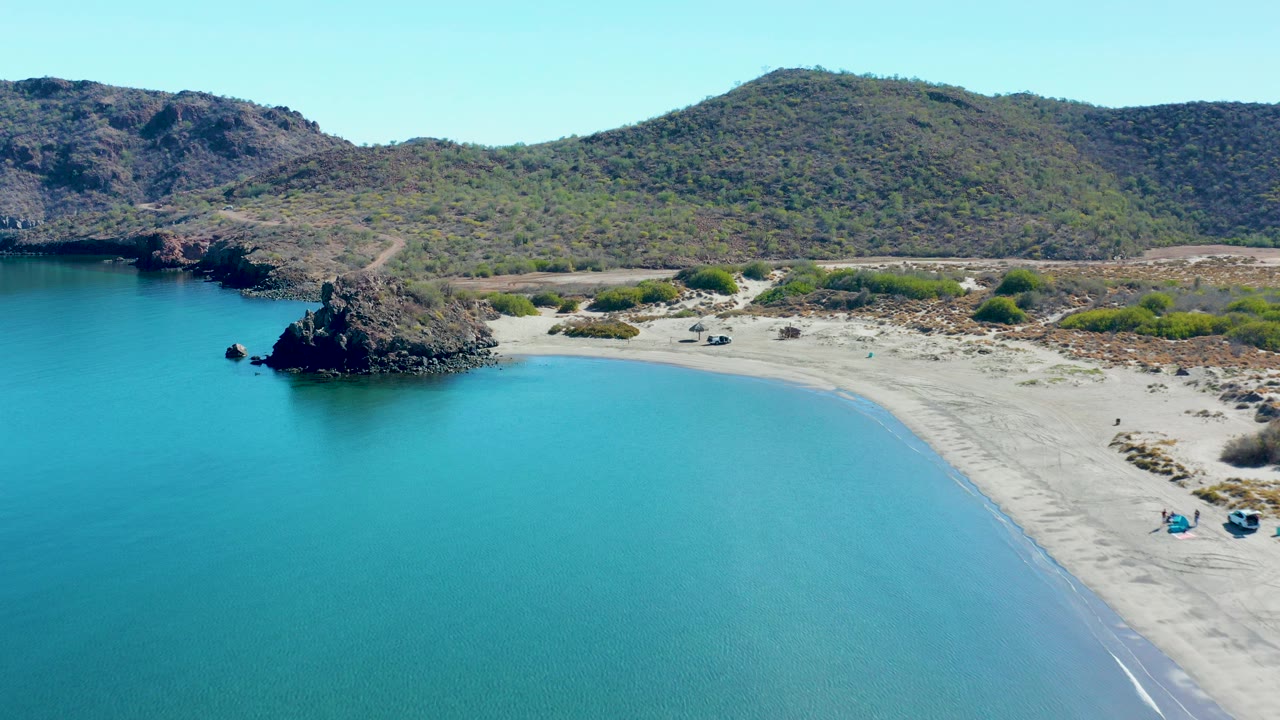 Aerial frame over Juncalito, Baja California Sur showing turquoise bay water, pale sand, a dark rocky point at left, scattered beach items, distant people, and low scrub hills under clear sky.