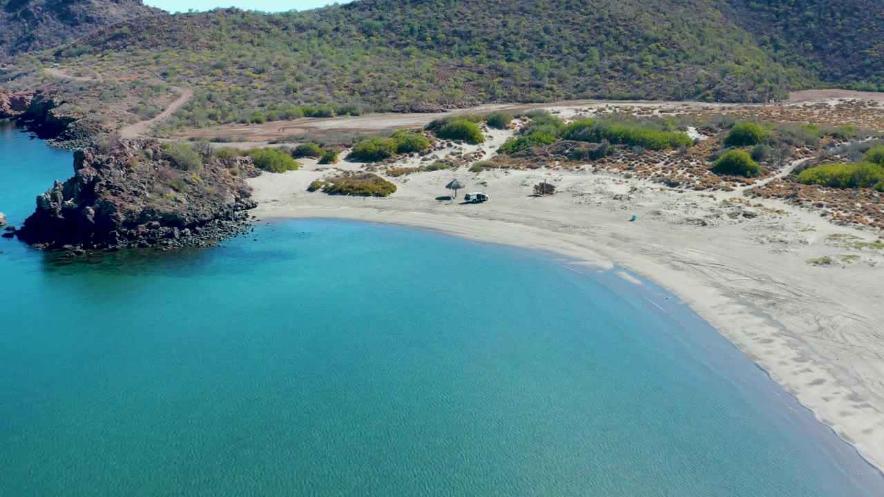 Aerial view over a Juncalito cove in Baja California Sur, with turquoise water in front, a pale sandy shore, a dark rocky outcrop at left, scrubby hills behind, and no visible people.