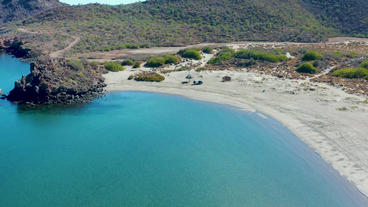 The Juncalito shoreline appears from above with shallow turquoise water, a clean crescent of pale sand, dark rocks at the left edge, green shrubs inland, and no visible people.