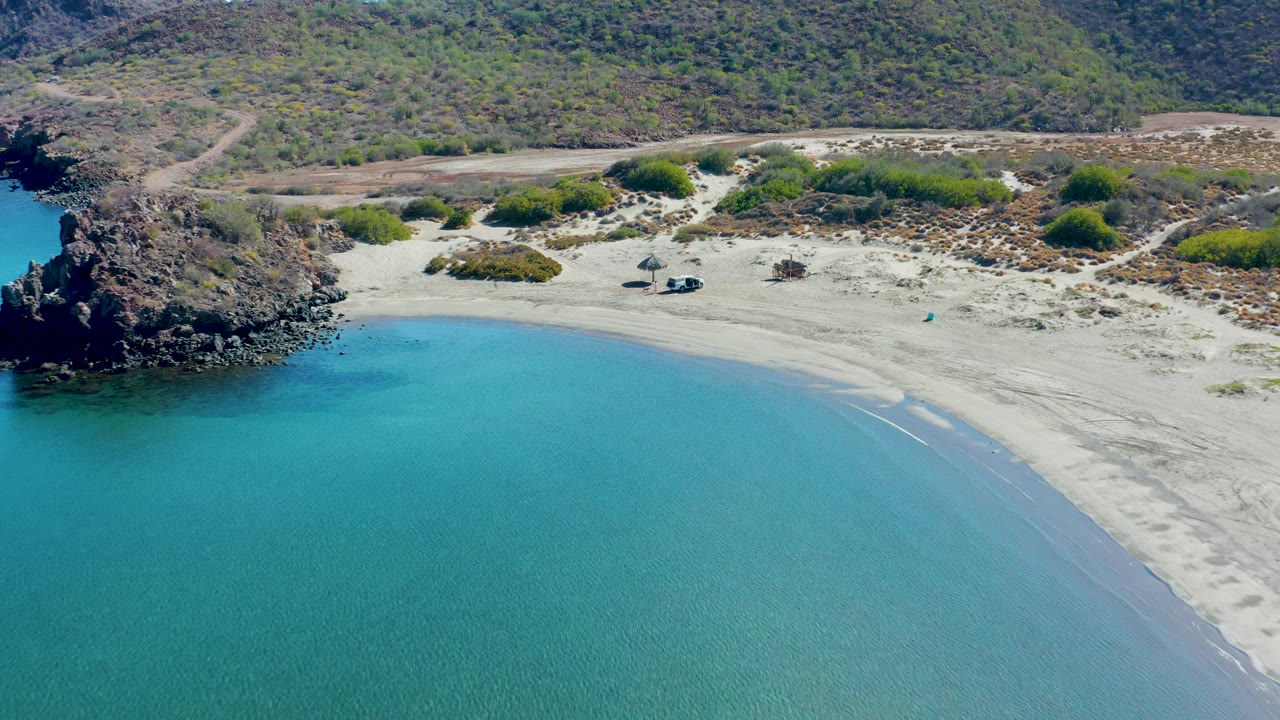 A wide aerial moment over Juncalito shows the calm turquoise cove, smooth pale beach, rugged dark rocks on the left, low coastal vegetation, hills in the background, and no visible people.