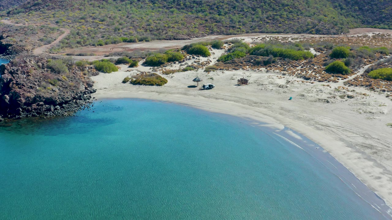 Aerial view past rocky shoreline toward empty sand, shrubs, and turquoise water.