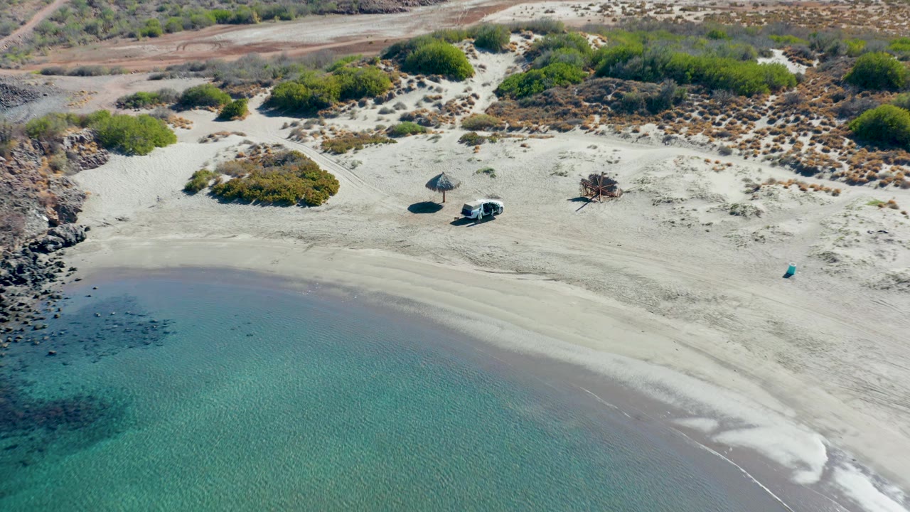High drone view in Juncalito, Baja California Sur, with the shoreline still prominent, the white van and thatched umbrella set back on pale sand, scrub vegetation behind them, and no visible people.