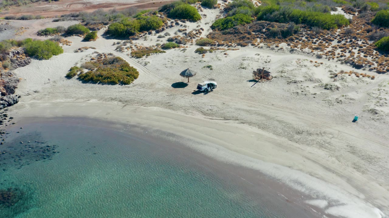 Drone frame over Juncalito, Baja California Sur, showing pale sand and shallow green water at left, a parked white van beside a thatched umbrella, brushy dunes in the background, and no visible people.