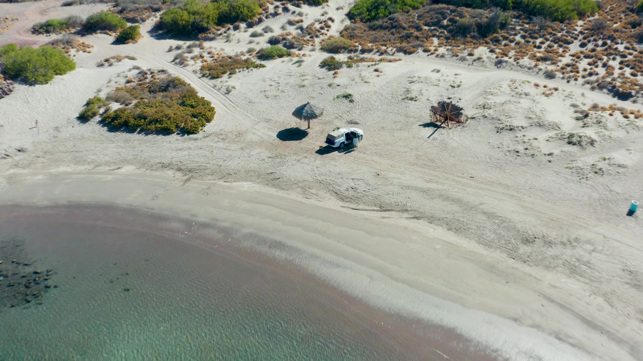 Closer aerial frame in Juncalito, Baja California Sur, with the clear water lower in frame, the white van and thatched umbrella more central on the sand, low shrubs above them, and no visible people.