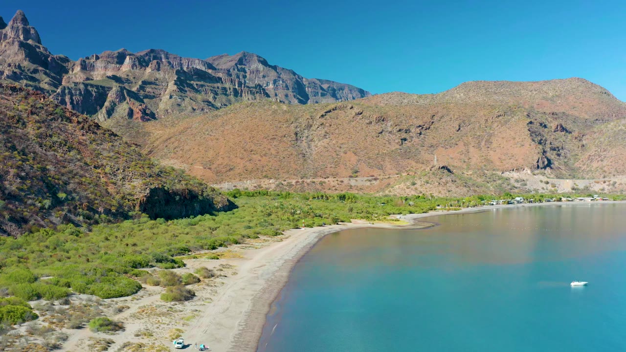 Aerial frame of Juncalito, Baja California Sur with the sandy shoreline slightly more centered, green scrub at the beach edge, calm blue water, distant mountains, and no visible people.