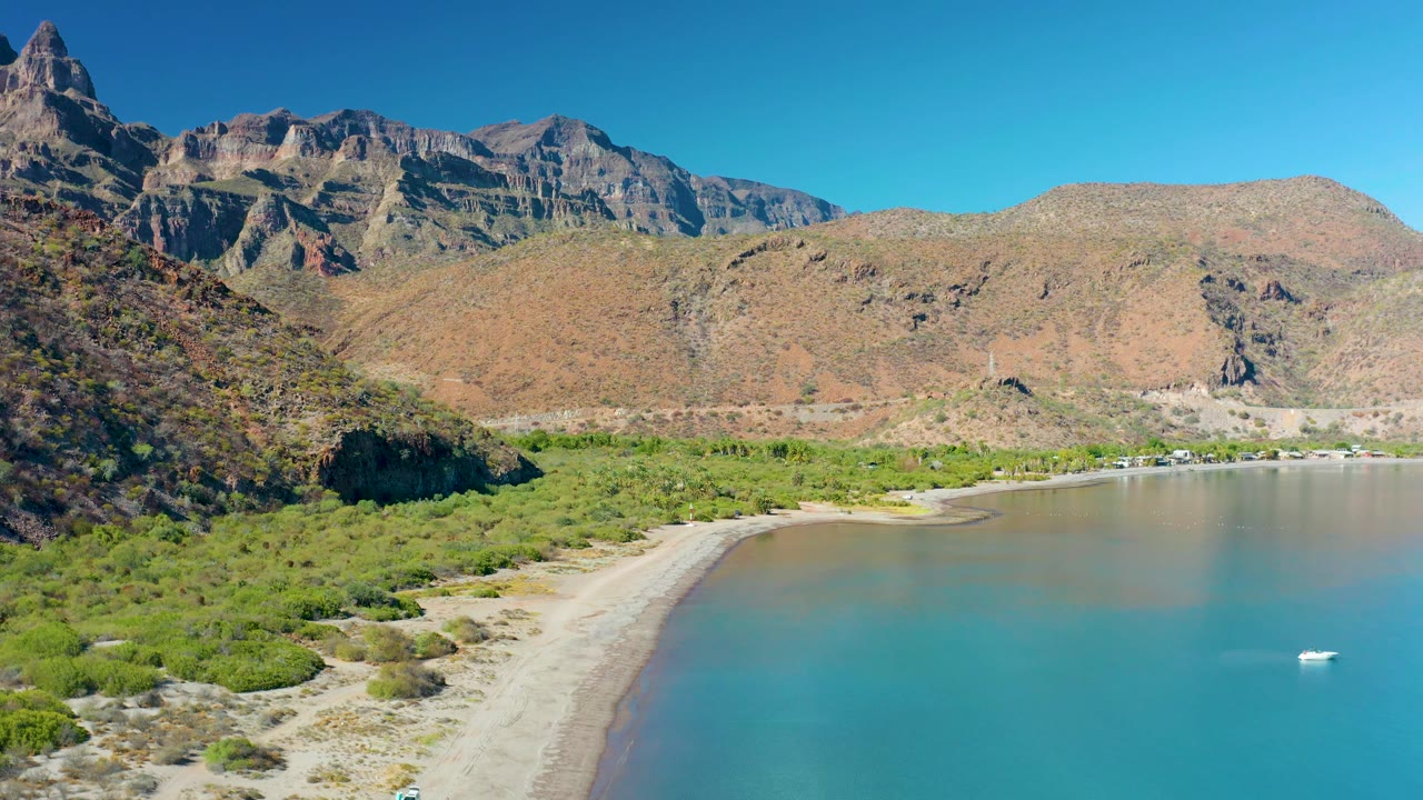 Aerial frame of Juncalito, Baja California Sur showing the boat farther right in the bay, a broad band of turquoise water, green coastal vegetation, dry mountains, and no visible people.
