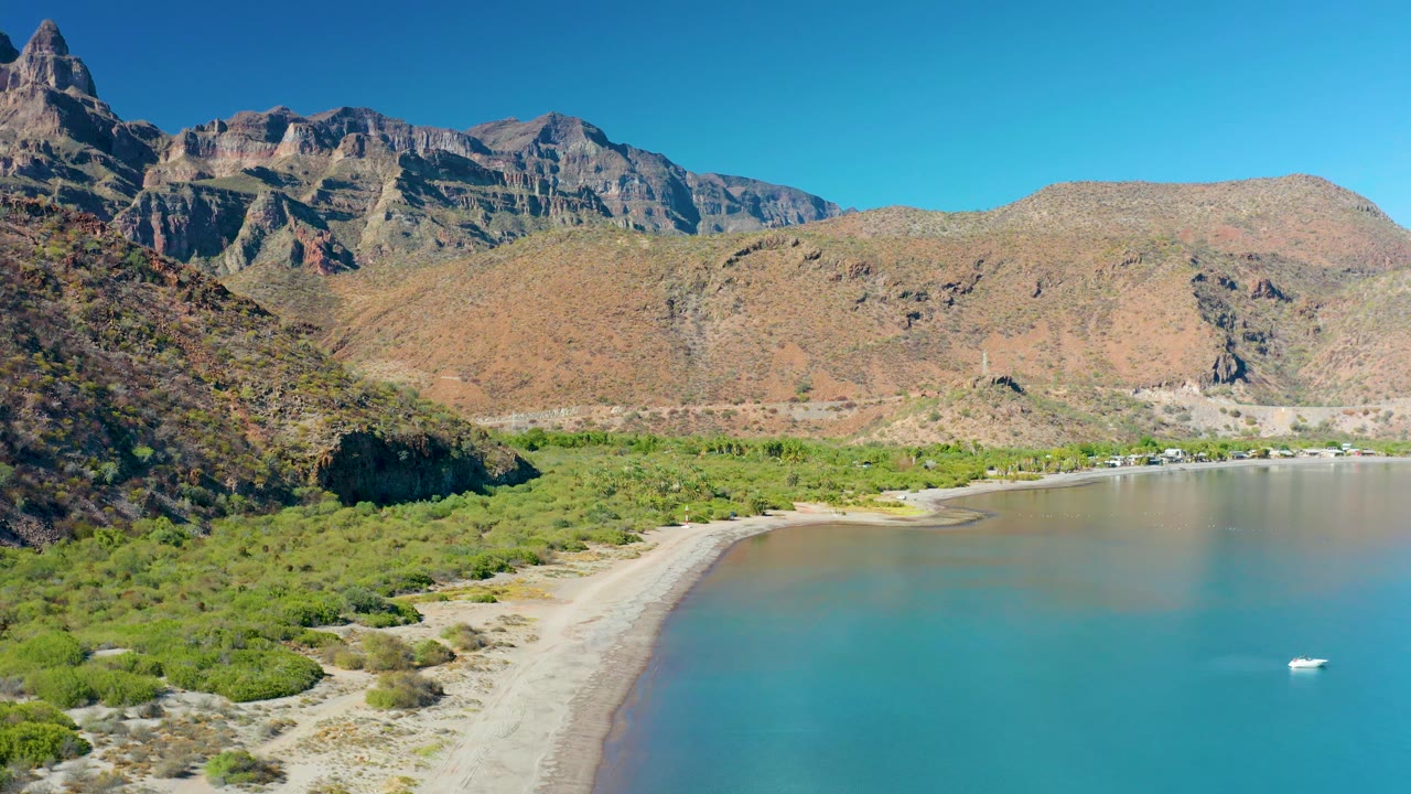 Aerial frame of Juncalito, Baja California Sur with more foreground vegetation visible, a pale sandy beach along the bay, muted brown shallows, rugged mountain ridges, and no visible people.