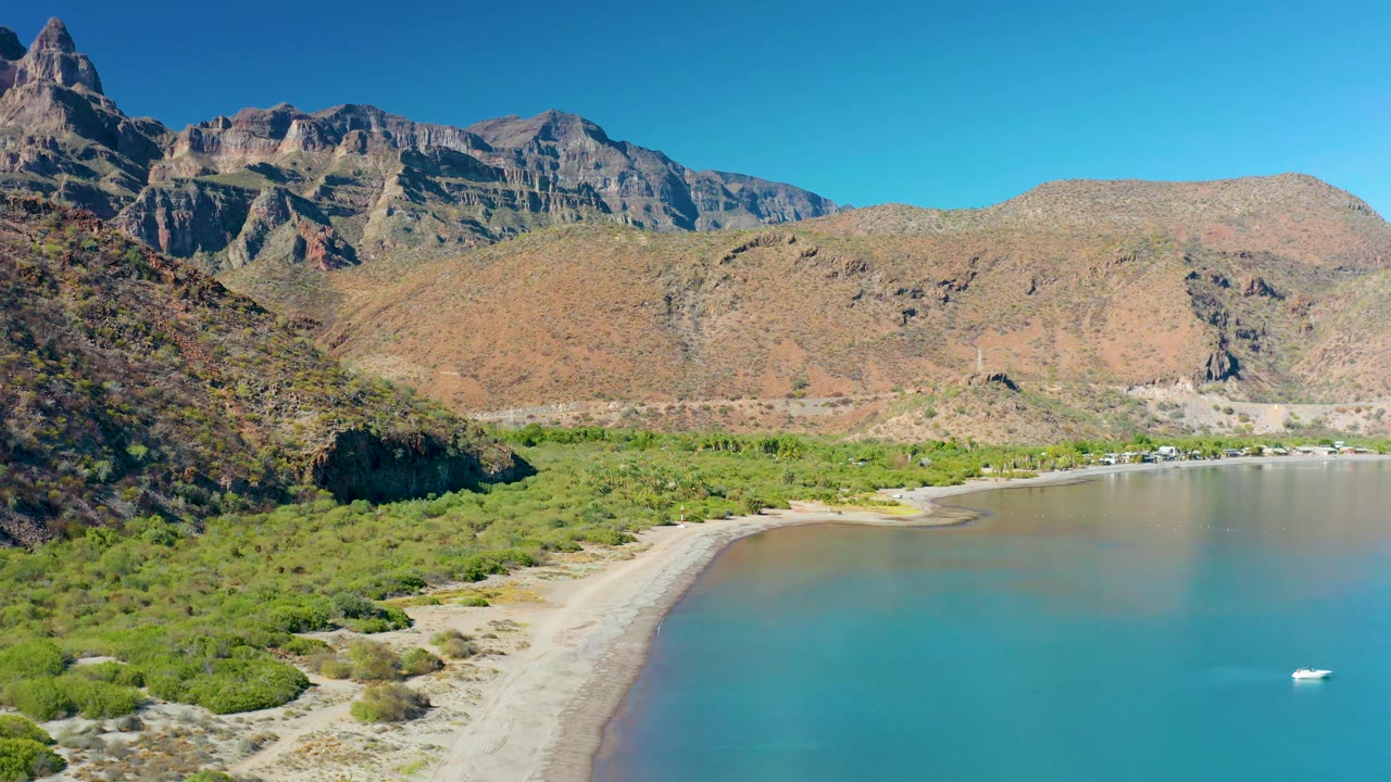 Aerial view in Juncalito, Baja California Sur, showing the shoreline slightly farther along the bay, with turquoise water, a white boat near the right edge, dry mountains, and no visible people.