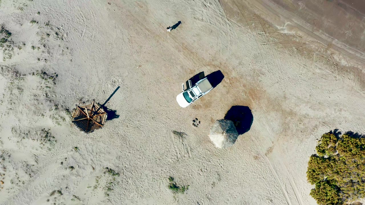 Overhead view in Juncalito, Baja California Sur showing a white car near sandy tracks, a wooden shade structure, sparse shrubs, and a small person near the upper frame.