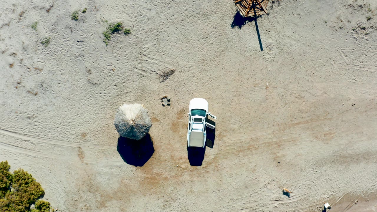 Overhead frame in Juncalito, Baja California Sur, Mexico, showing a white car beside a straw umbrella on pale sandy terrain with hard shadows and no visible people.