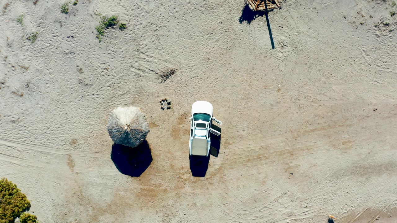 Top-down frame from Juncalito showing the parked white car next to the umbrella, with scattered vegetation, faint vehicle tracks, and no visible people.