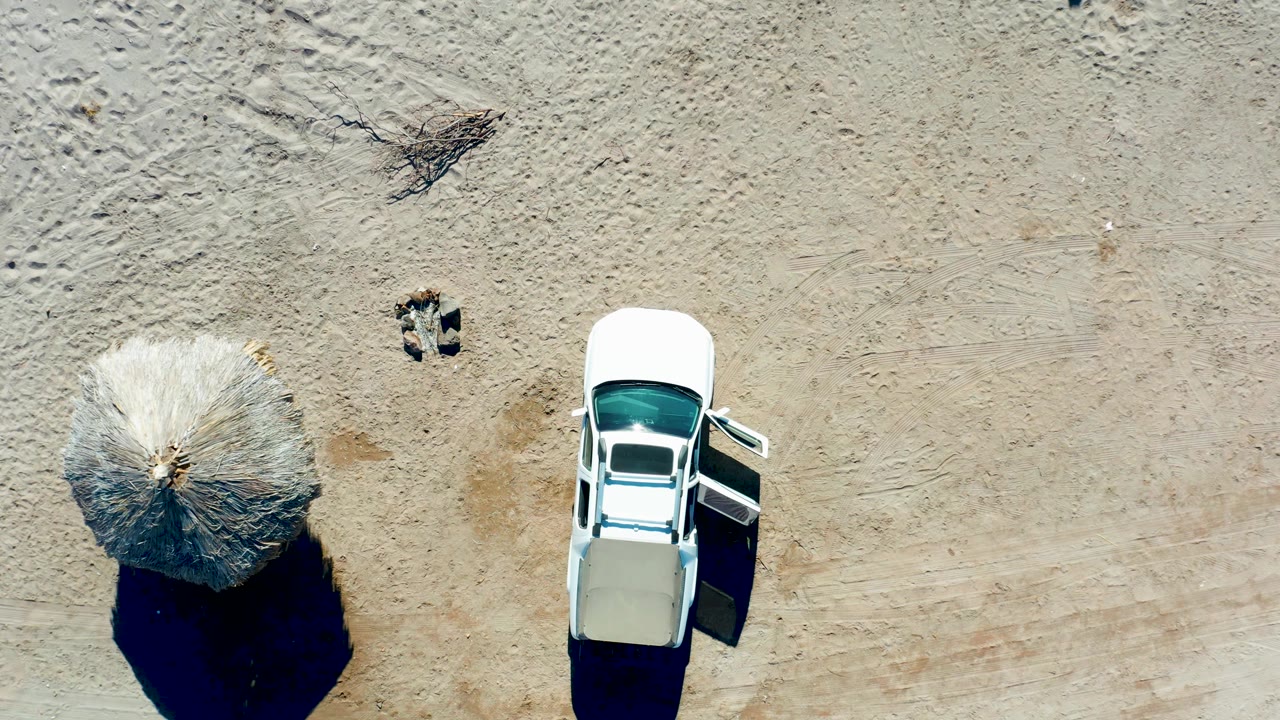 Top-down Juncalito frame with the white car shifted slightly right of center, the thatched umbrella on the left, visible tire marks, and no visible people.