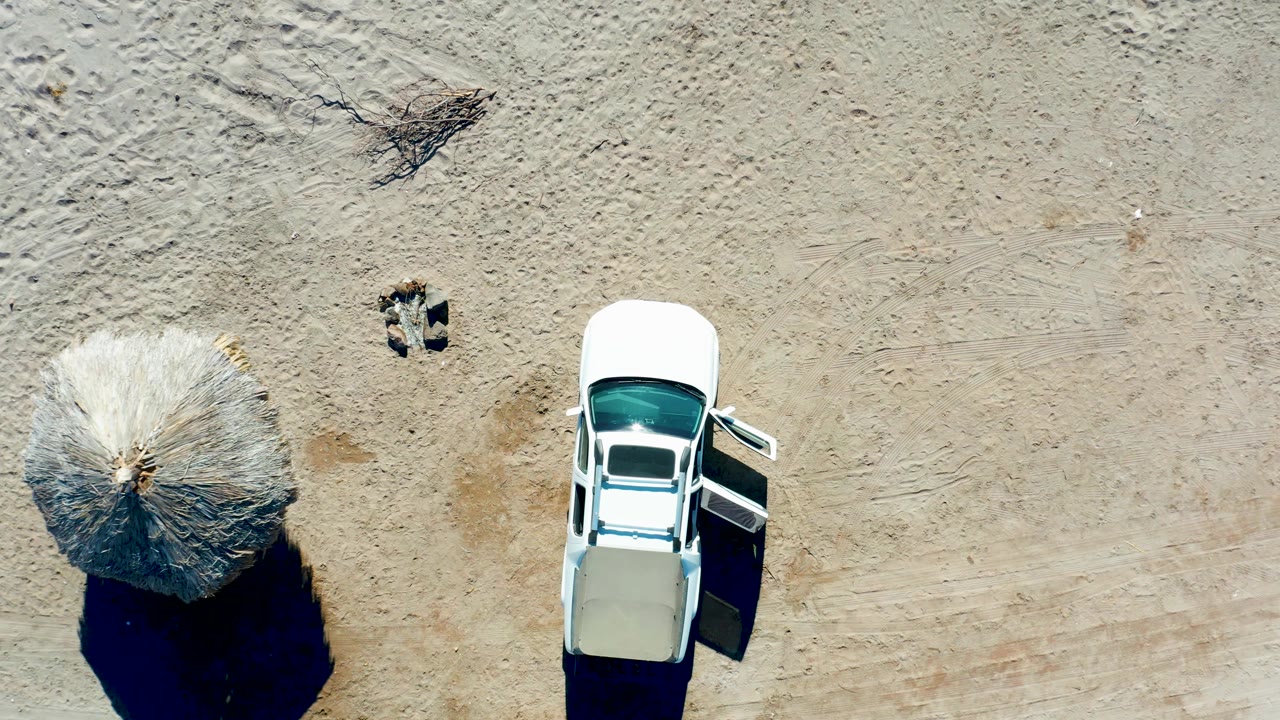 Overhead frame in Juncalito, Baja California Sur with the white car near the right edge, a round thatched umbrella left of it, sand tracks, and no visible people.