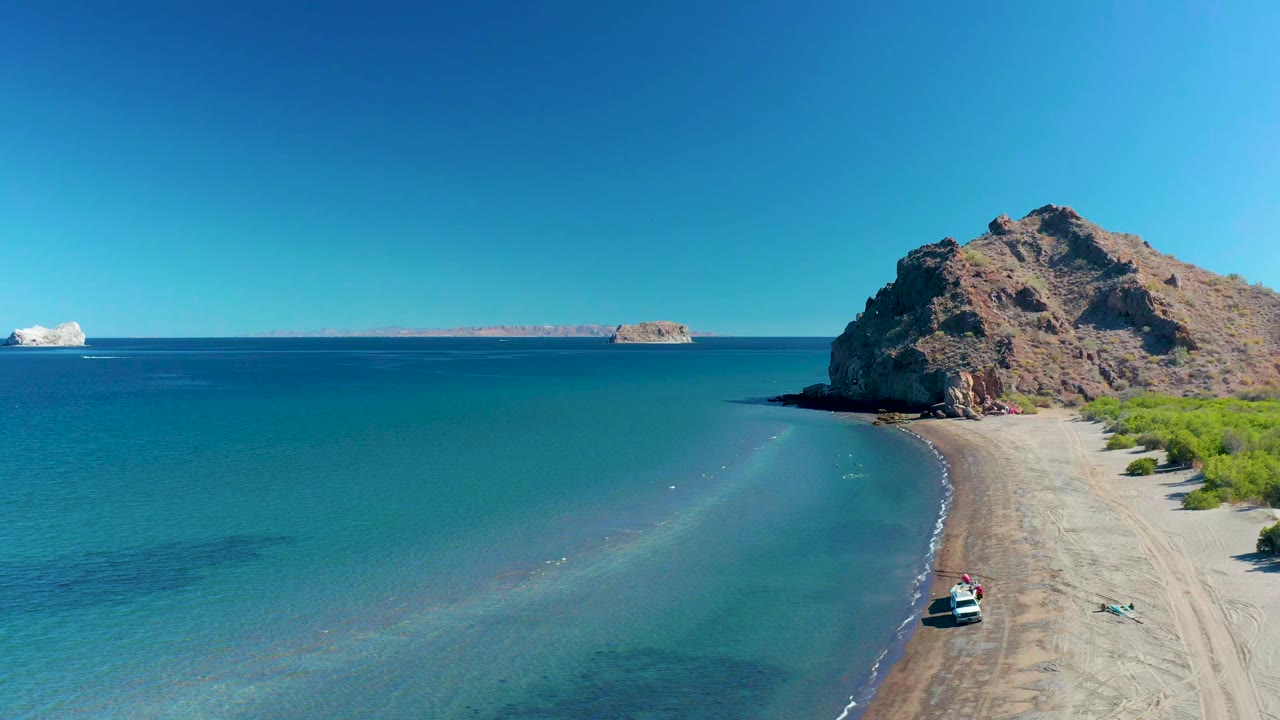 Aerial view in Ligui, Baja California Sur with the small boat slightly farther along the sandy shoreline, calm blue water filling the left side, a rocky cliff at right, and no visible people.