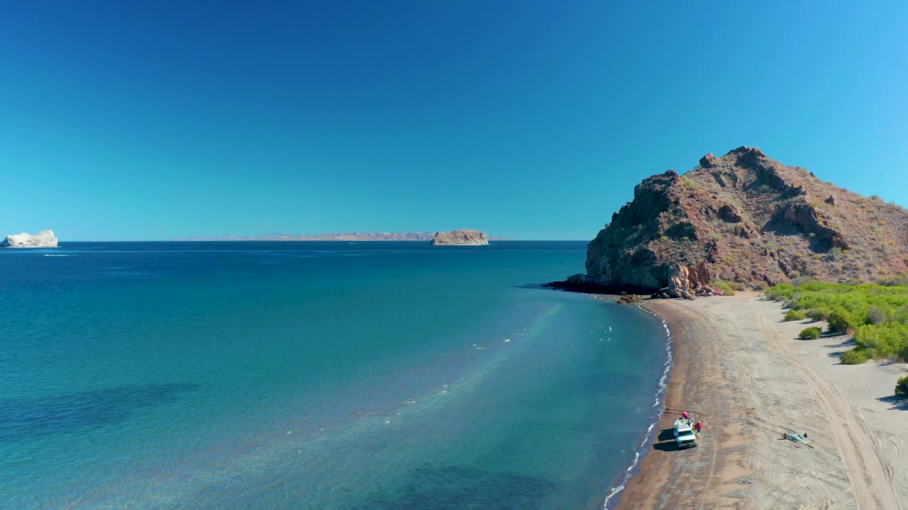 Ligui, Baja California Sur appears from above with more open water visible on the left, the sandy shoreline bending toward the rocky cliff, a small boat near the lower right, and no visible people.