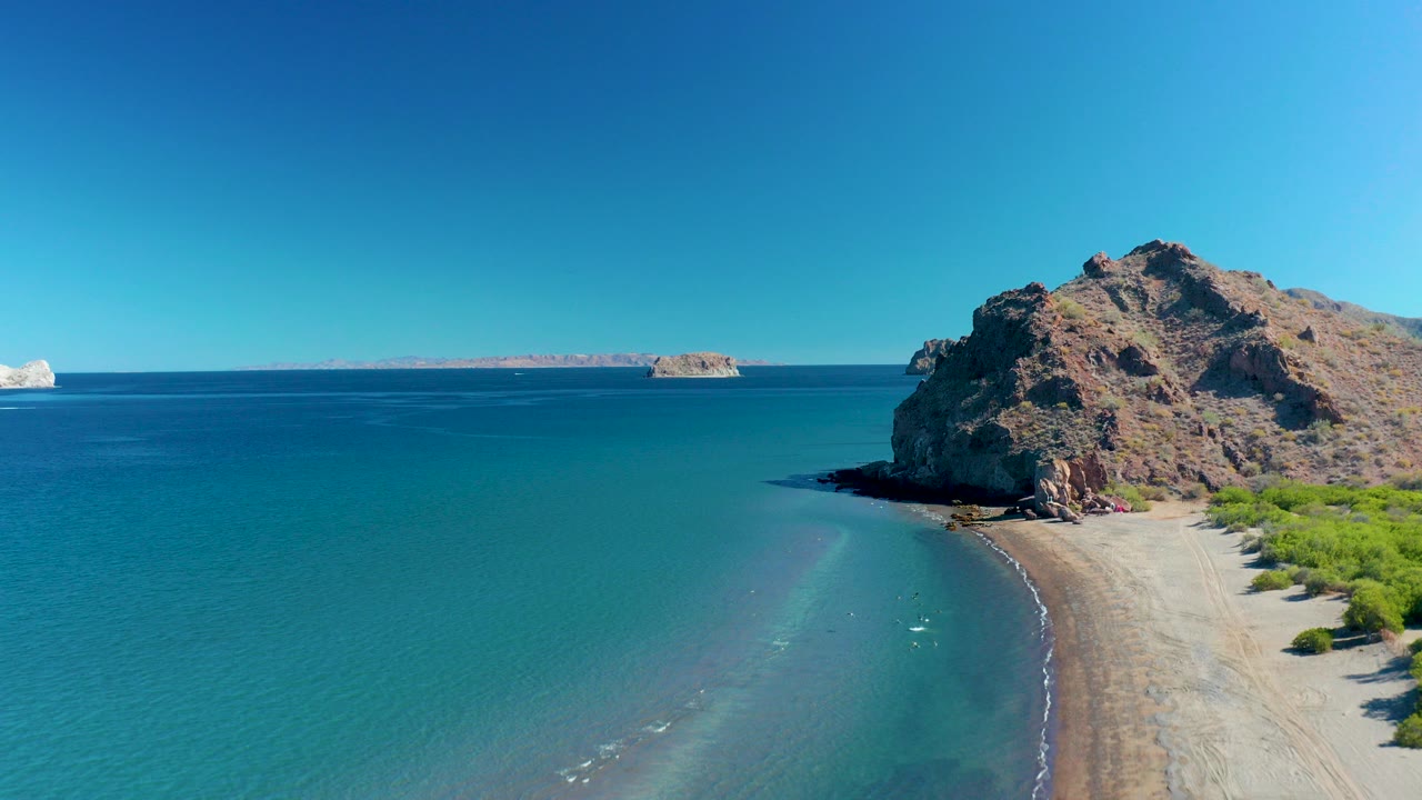 Aerial frame over Ligui, Baja California Sur, Mexico showing blue ocean in the foreground, a curved sandy shore beside a rocky cliff on the right, distant islands on the horizon, and no visible people.