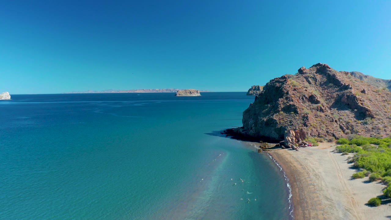 Aerial view at 0:03 in Ligui with more open water visible to the left, the rocky point and sandy shore on the right, a small distant island near the horizon, and no visible people.