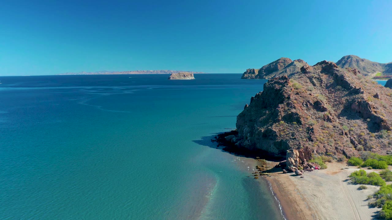 Aerial frame at 0:01 in Ligui, Baja California Sur, with the rocky cliff still dominant on the right, green-blue shallows below, a thin beach edge, distant landforms, and no visible people.