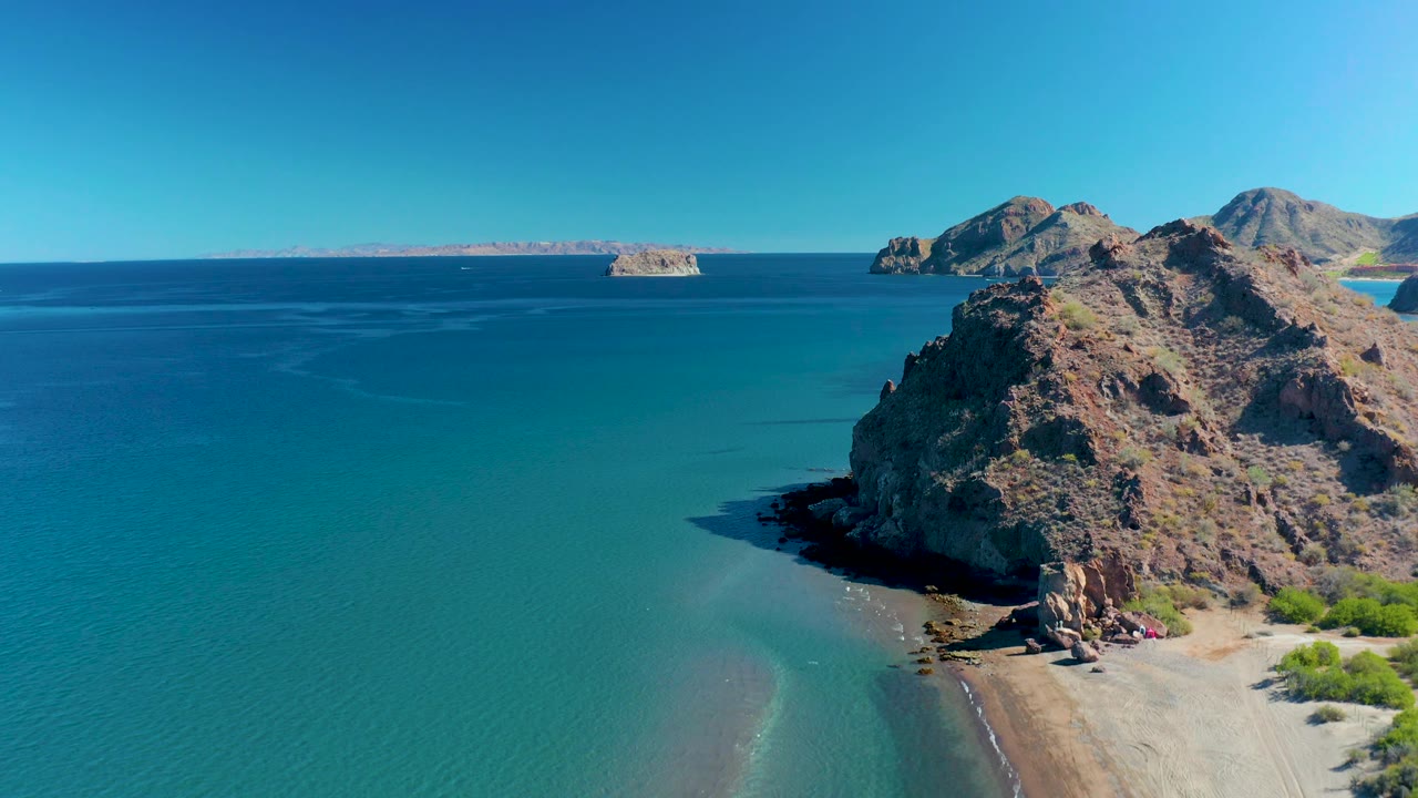 Aerial frame at 0:02 over Ligui, Baja California Sur, showing the headland slightly farther right in frame, calm turquoise water across the foreground, pale sand at the cliff base, and no visible people.