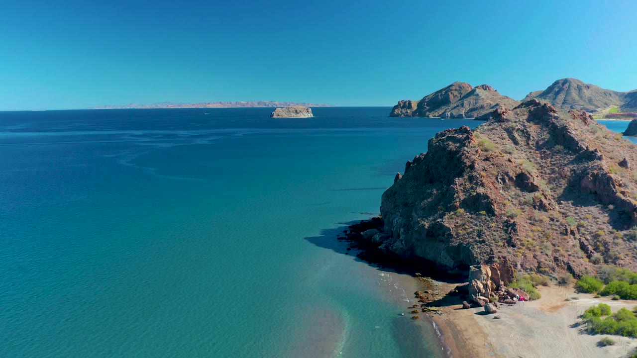 Aerial frame at 0:03 in Ligui, Baja California Sur, showing more open water to the left, the rocky point and small beach on the right, distant islands near the horizon, and no visible people.